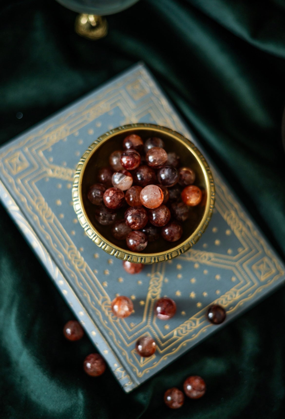 Feuerquarz Mini-Sphären in goldener Schale auf blau-goldenem Buch, umgeben von dunklem grünen Samt – magische Kristalle für Kreativität und innere Kraft.